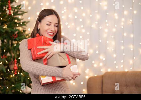 Glückliche junge Frau hält viele Geschenkboxen mit Weihnachtsbaum Und Lichter im Hintergrund Stockfoto