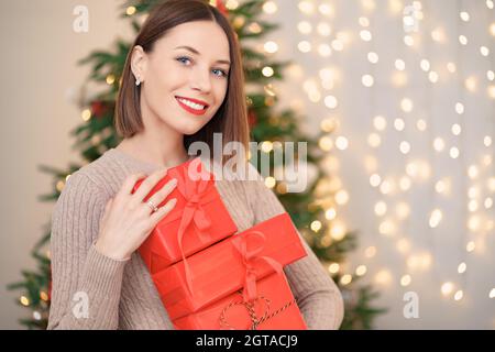 Glückliche junge Frau hält viele Geschenkboxen mit Weihnachtsbaum Und Lichter im Hintergrund Stockfoto