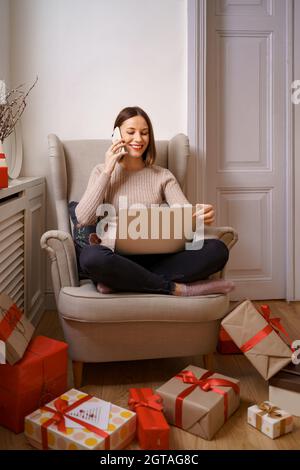 Schöne junge Frau mit Laptop im Gespräch auf Handy, während Sitzen in einem bequemen Sessel umgeben von Geschenken zu Hause Stockfoto