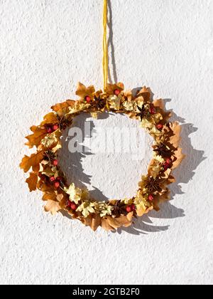 Getrockneter Blumenkranz aus trockenen Herbstblättern und Beeren hängen an der weißen Wand mit strukturierter Tapete auf gelber Kordel. Stockfoto