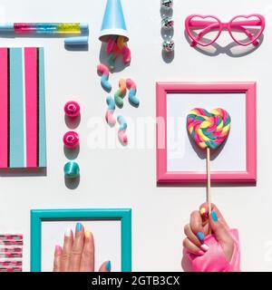 Sammlung von Party-Objekten und Süßigkeiten, Draufsicht, flaches Lay. Schokolade, Süßigkeiten. Notizbuch und Regenbogen-Lolly knallen in der Hand. Festliche geometrische Anordnung in Stockfoto