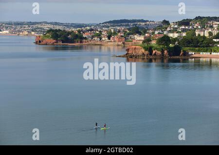 Stand-up-Paddlebarder in Tor Bay, mit Blick auf Corbyn's Head und Livermead von der Torquay-Küste aus. Stockfoto