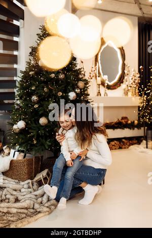 Portrait einer schönen Frau, die sich an der kleinen, kleinen, Backentochter küsst, die neben einem geschmückten Weihnachtsbaum sitzt. Liebevolle Mutter umarmt entzückendes Kind Stockfoto