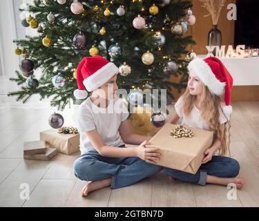 Kinder - ein Junge und ein Mädchen spielen in der Nähe des Weihnachtsbaums. Wohnzimmer mit Weihnachtsbaum und Dekorationen. Neujahr. Geschenk geben. Stockfoto