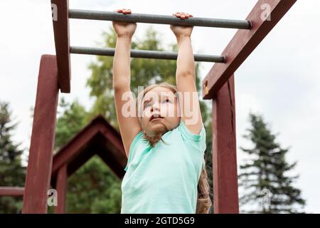 Ein aktives junges Mädchen, fit sportliche Kinderklettern, Outdoor-Bewegung, hängen an einer Bar auf einem Spielplatz, Porträt, Nahaufnahme. Kinder und gesunde körperliche Stockfoto