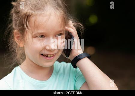 Ein fröhliches, fröhliches kleines Kind, ein kleines Mädchen, das auf ihrem Mobiltelefon redet und es in der Hand neben dem Ohr lächelt, ein schlaues Band trägt und lacht. Kid po Stockfoto