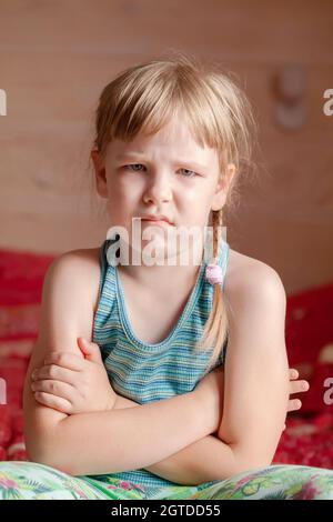 Ein einziges beleidigtes Kind, kleines Mädchen im Schulalter, das zu Hause mit gekreuzten Armen auf dem Bett sitzt, unglücklich, rebellisches Kind einfaches vertikales Porträt, Stockfoto