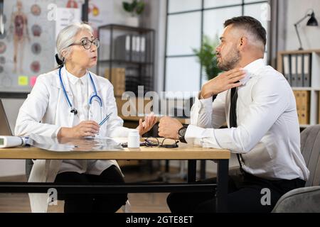 Kaukasischer Mann in Geschäftskleidung, der sich während des Termins in einer modernen Klinik über Halsschmerzen beschwert. Reife Ärztin in Brillen und Uniform, die am Schreibtisch sitzt und aufmerksam zuhört, männlicher Patient. Stockfoto