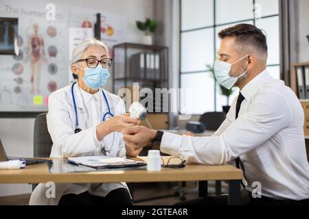 Medizinische Mitarbeiterin mit modernem Infrarot-Thermometer zur Temperaturkontrolle des kaukasischen Geschäftsmannes. Arzt und Patient tragen eine schützende Gesichtsmaske. Kabinett der modernen Klinik. Stockfoto
