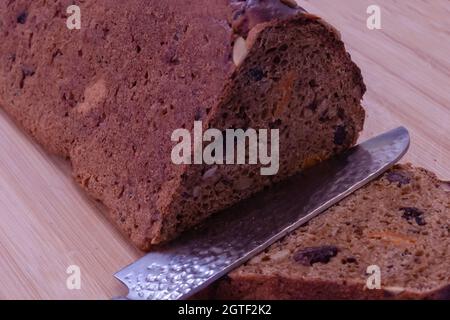 Brot mit einem Messer auf einem hellen Holzbrett schneiden, Nüsse, getrocknete Aprikosen und Rosinen sind im Brot sichtbar Stockfoto