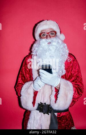 Mann, der als Weihnachtsmann gekleidet war, mit einem Smartphone mit einer lustigen Brille Stockfoto