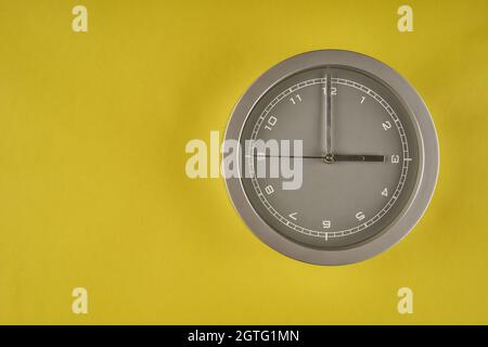 Graue, runde Bürouhr, die auf einem gelben Hintergrund isoliert auf drei Uhr zeigt Stockfoto