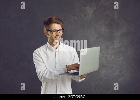 Lächelnder Mann steht auf grauem Hintergrund und gibt Text auf Laptop ein, den er in die Hände hält. Stockfoto