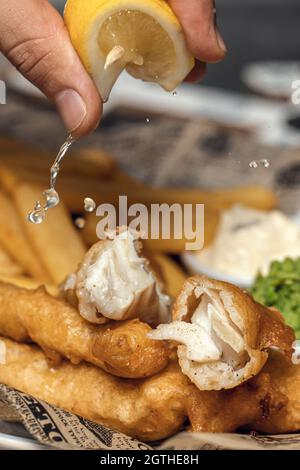 Traditionell britischer knuspriger Fisch und Chips mit pürierten Erbsen, Tatarensauce auf Zeitung, Zitrone auf Fisch und Chips quetschen Stockfoto