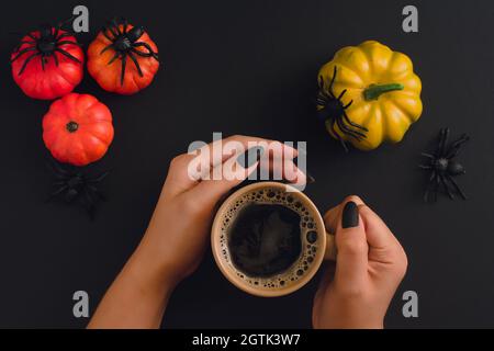 Schwarze, gepflegte Hände halten Cappuccino-Becher auf Halloween-Hintergrund mit devotiven Kürbissen und Spinnen. Blick von oben. Stockfoto