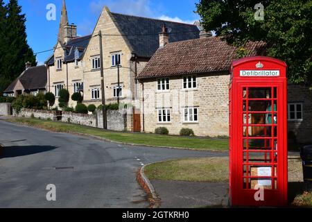 Eine alte rote Telefonbox, die für einen Defibrillator umfunktioniert wurde, neben dem Village Green in Barnwell, Northamptonshire, Großbritannien Stockfoto