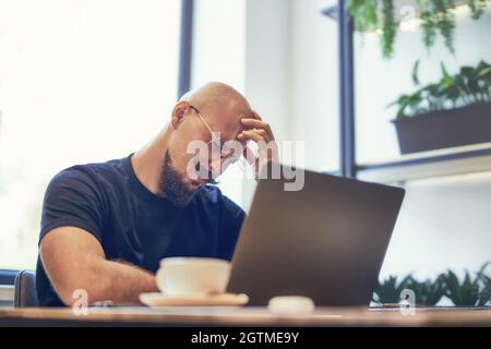 Unglücklicher Mann legte seine Hand auf den Kopf, müder Geschäftsmann sitzt mit Laptop am Schreibtisch und hat Kopfschmerzen Stockfoto