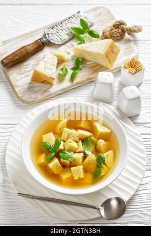 Zuppa imperiale, italienische Suppe aus Hühnerbrühe mit Parmesan-Eierknödeln, bestreut mit geriebenem Käse und Basilikum in einer weißen Schüssel auf einem Holztisch,V Stockfoto
