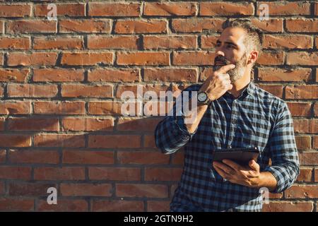 Porträt eines modernen Geschäftsmannes mit Bartdenken und mit einem digitalen Tablet, während er im Freien vor einer Backsteinmauer steht. Stockfoto
