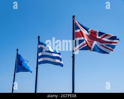Symbol für Griechenland, Großbritannien und die EU. Flaggen des Vereinigten Königreichs, Griechenlands und der Europäischen Union auf Fahnenmasten, die im Wind winken. Klarer blauer Himmel, sonniger Frühlingstag. Stockfoto