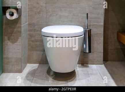 Moderne Toilette Inneneinrichtung, hängende weiße Toilettenschüssel Papierrollenhalter und Bürste. Luxuriöse Badezimmerausstattung an Wänden und Bodenfliesen aus Marmor Stockfoto