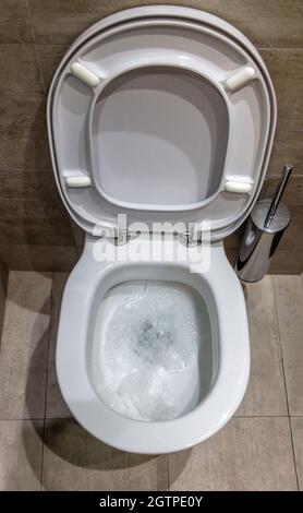 Toilettenschüssel spült Wasser, über Ansicht. Weiße Toilettenschüssel mit offener Abdeckung und Bürste. Luxuriöse Badezimmerausstattung an Marmorwänden und Bodenbeleuchtung Stockfoto