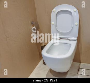 WC-Innenausstattung, weiße Keramikschale mit offenem Deckel und schwarzer Bürste. Moderne Badezimmerausstattung an braunen Wänden und gefliestem Boden. Stockfoto