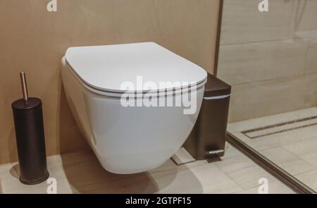 WC-Innenausstattung, weiße Keramikschale und schwarze Bürste. Moderne Badezimmerausstattung an braunen Wänden und gefliestem Boden. Stockfoto