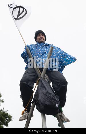 Farnborough, Großbritannien. Oktober 2021. Ein Klimaaktivist der Extinction Rebellion sitzt auf einem Stahlstativ verriegelt, um den Eingang zum Flughafen Farnborough zu blockieren. Aktivisten blockierten drei Zugänge zum privaten Flughafen, um erhöhte Kohlendioxidwerte hervorzuheben, die von superreichen Passagieren mit privaten Jets und "Greenwashing" vom Flughafen produziert werden, indem sie einen Wechsel zu nachhaltigem Flugkraftstoff (SAF) ankündigten. Kredit: Mark Kerrison/Alamy Live Nachrichten Stockfoto