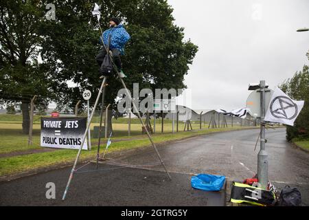 Farnborough, Großbritannien. Oktober 2021. Ein Klimaaktivist des Extinction Rebellion ist auf einem Stahlstativ befestigt, um den Eingang zum Flughafen Farnborough zu blockieren. Aktivisten blockierten drei Zugänge zum privaten Flughafen, um erhöhte Kohlendioxidwerte hervorzuheben, die von superreichen Passagieren mit privaten Jets und "Greenwashing" vom Flughafen produziert werden, indem sie einen Wechsel zu nachhaltigem Flugkraftstoff (SAF) ankündigten. Kredit: Mark Kerrison/Alamy Live Nachrichten Stockfoto