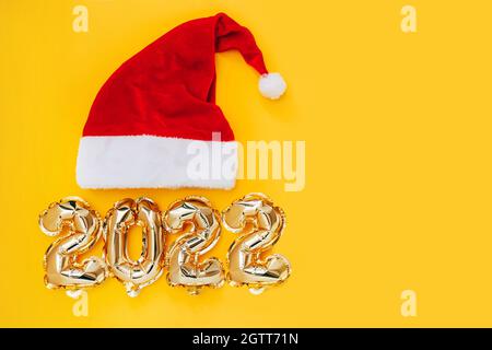 Folienballons in Form von Zahlen 2022 und Weihnachtsmütze auf gelbem Hintergrund. Neujahr. Stockfoto