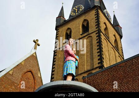 Lebensechte Säulenheilige Skulptur einer Mutter mit ihrem Kind vom Künstler Christoph Pöggeler. St. Lambertus Kirche im Hintergrund. Stockfoto