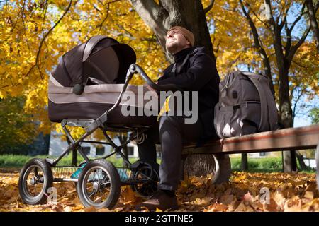 Glücklicher junger Vater mit Kinderwagen sitzt auf der Bank in der Natur im Park Stockfoto