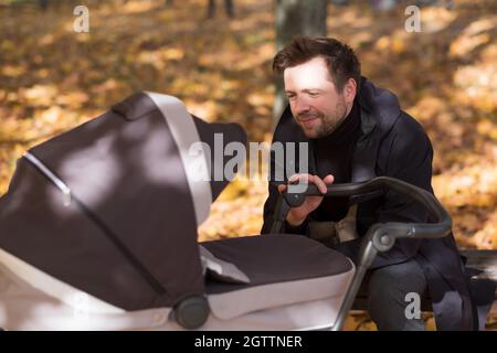 Glücklicher junger Vater mit Kinderwagen sitzt auf der Bank in der Natur im Park Stockfoto