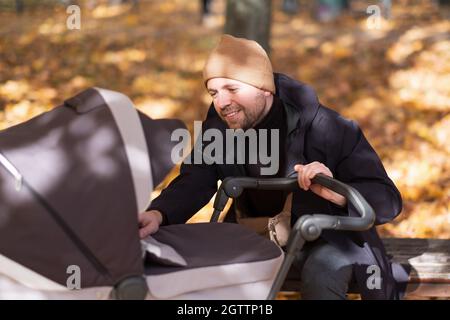 Glücklicher junger Vater mit Kinderwagen sitzt auf der Bank in der Natur im Park Stockfoto