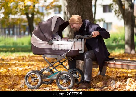 Glücklicher junger Vater mit Kinderwagen sitzt auf der Bank in der Natur im Park Stockfoto