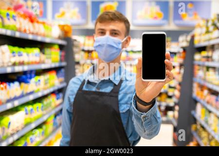 Junger Mann im Geschäft mit einer chirurgischen medizinischen Maske in schwarzer Schürze, die das Smartphone-Display im Hintergrund des Marktes zeigt. Stockfoto