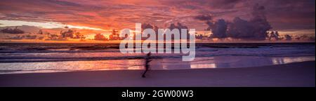 Bewegungsunschärfe des Joggers, der bei Sonnenaufgang am Meeresstrand läuft, mit orangefarbenem und purpurem Sonnenaufgang und wolkenverdunkelter Himmel, der von den Wellen reflektiert wird Stockfoto