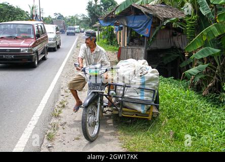 Ein älterer Mann auf einem Motorrad mit einem Seitenwagen, der mit großen Säcken gefüllt ist, an einer befahrenen Straße zwischen Padang an Bukittinggi, West-Sumatra, Indonesien. Stockfoto