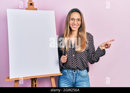 Schöne hispanische Frau steht bei Maler Staffelstand halten Pinsel lächelnd glücklich zeigen mit Hand und Finger zur Seite Stockfoto