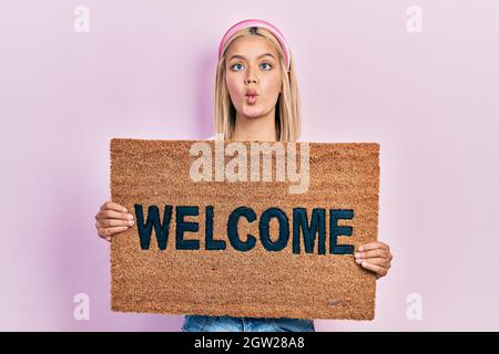 Schöne blonde Frau hält willkommen Fußmatte machen Fisch Gesicht mit Mund und schielende Augen, verrückt und komisch. Stockfoto