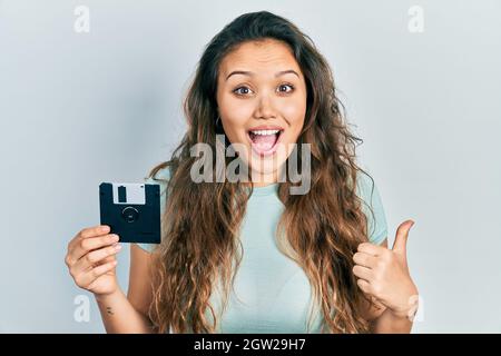 Junge hispanische Mädchen hält Diskette mit dem Daumen nach oben auf die Seite lächelnd glücklich mit offenem Mund Stockfoto