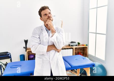 Hübscher junger Mann, der in der Schmerzlinik arbeitet, mit der Hand auf dem Kinn, der über Fragen nachdenkt, über nachdenklichen Ausdruck. Lächelnd mit nachdenklichem Gesicht. Zweifel Stockfoto
