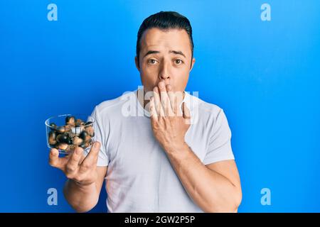 Hübscher junger Mann mit rohen Haselnüssen, die den Mund mit der Hand bedecken, schockiert und vor Fehlern fürchtend. Überraschter Ausdruck Stockfoto