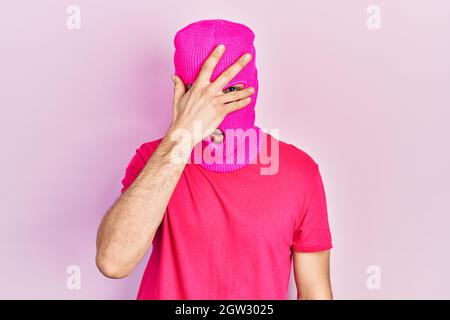 Junger hispanischer Mann mit modern gefärbten Haaren trägt rosa Balaclava Maske Gesicht schauend in Schock über Gesicht und Augen mit der Hand, Blick durch die Finger ein Stockfoto