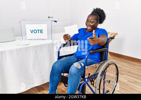 Junge afrikanische Frau sitzt auf Rollstuhl Abstimmung Putting umhüllen in Wahlurne zeigt Finger zur Kamera mit glücklichen und lustigen Gesicht. Gute Energie und V Stockfoto