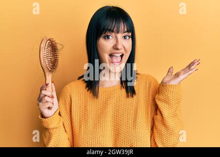 Junge hispanische Frau, die den Kamm hält und Haare verliert, feiert den Sieg mit einem glücklichen Lächeln und einem Siegerausdruck mit erhobenen Händen Stockfoto