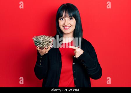 Junge hispanische Frau hält Sonnenblumenkerne Schüssel lächelnd glücklich zeigt mit Hand und Finger Stockfoto