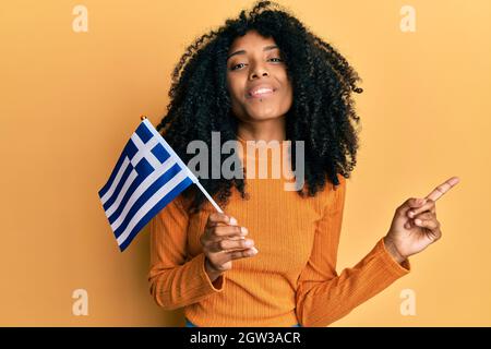 afroamerikanische Frau mit afrohaarigen Händen, die die griechische Flagge lächelnd hält und mit Hand und Finger zur Seite zeigt Stockfoto