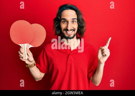 Junger hispanischer Mann mit roten Ping-Pong-Schlägern, lächelnd, zeigt glücklich mit Hand und Finger zur Seite Stockfoto